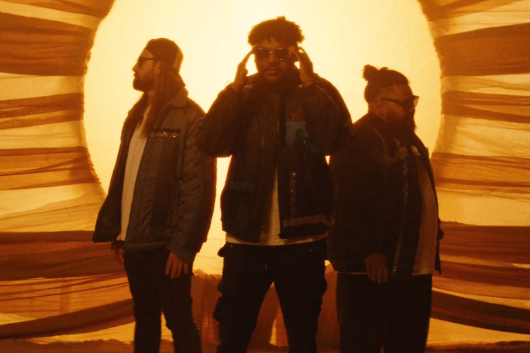 First Nations hip-hop group DEM MOB standing together silhouetted against a bright glowing circle backlighting draped fabric in warm golden hues.